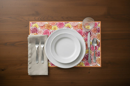 Set table with white plates, silverware, and a colorful placemat with citrus designs on a wooden surface