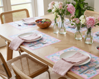 Dining table set with plates, napkins, and a floral arrangement in a bright room.