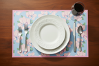Table setting with a floral placemat, white plate, silverware, and glass on a wooden table.