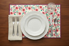 Dining table setting with floral placemat, white plates, silverware, and a glass on a wooden surface.