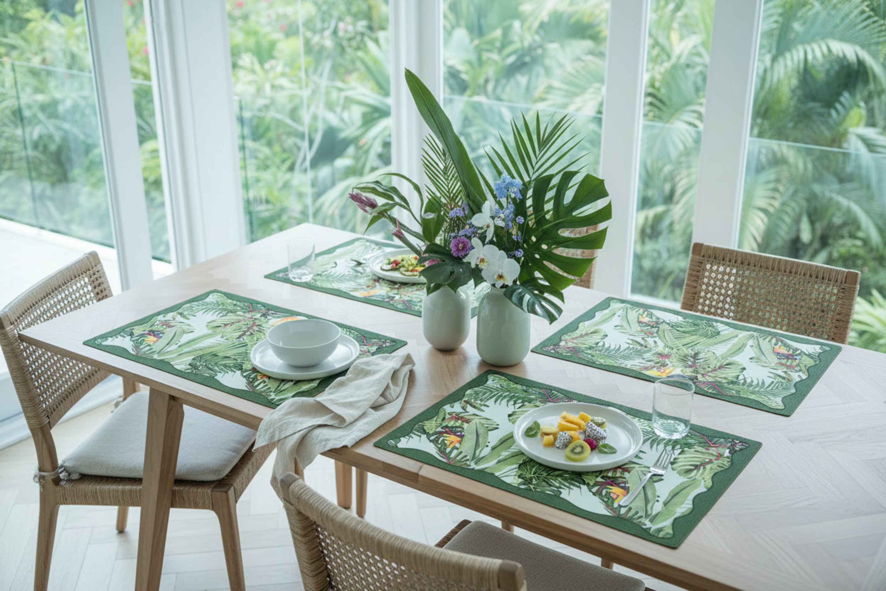 Dining table set with green placemats, white plates, and a vase of flowers in a bright room with large windows.