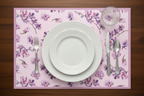 Table setting with floral placemat, white plates, silverware, and a glass on a wooden surface.