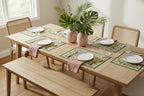 Pattern of toucans and monstera leaves on a green leafy background printed on a paper placemat set on a wooden dining table with plates and golden color silverware