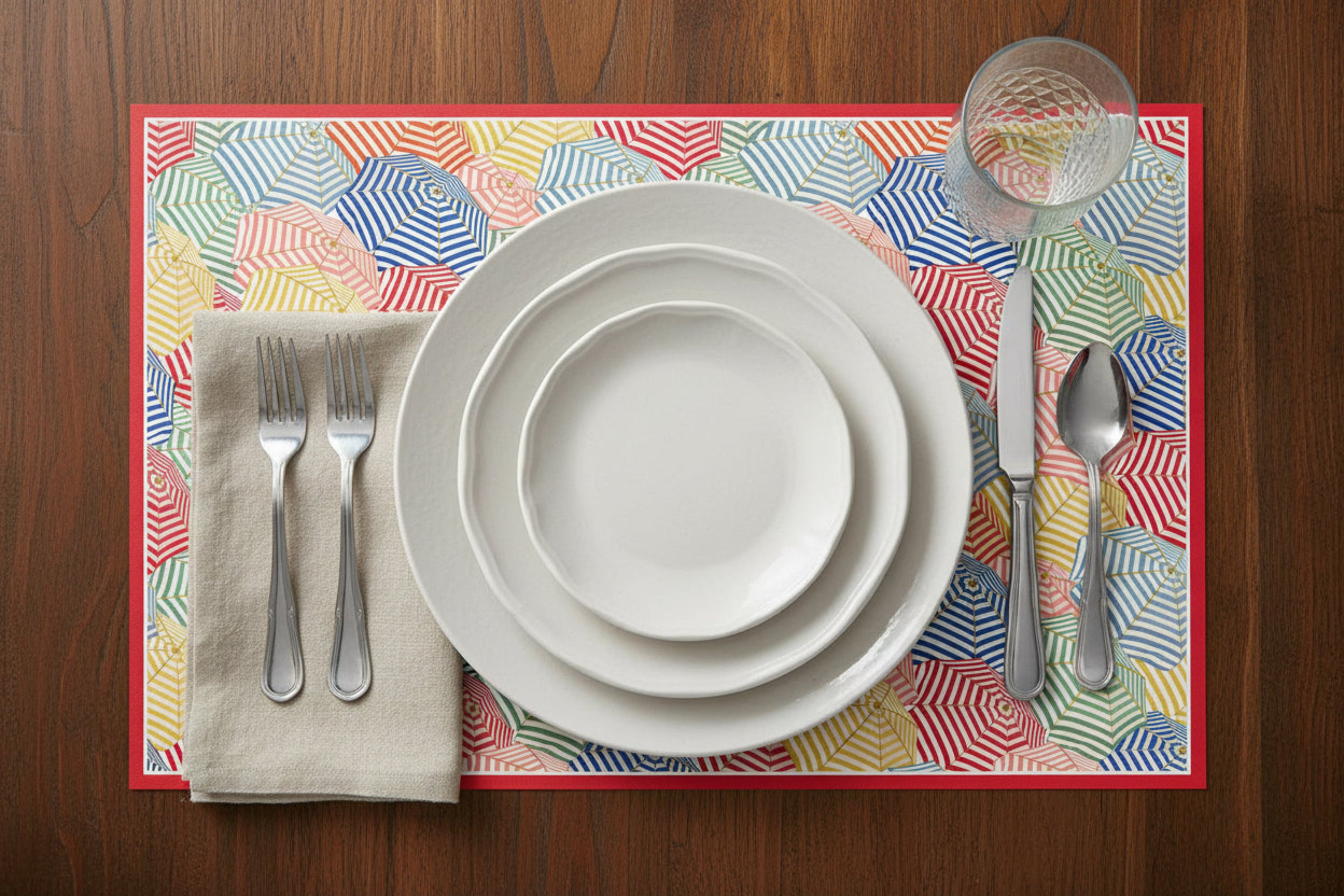 Dinnerware set on a colorful umbrella designs placemat with cutlery on a wooden table.